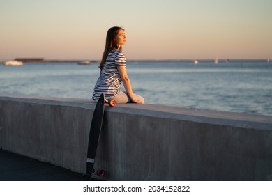 Teen skater girl sit at sidewalk during sunset at seaside. Cute young woman enjoy summer evening at river or sea looking at sun setting in water. Urban female with longboard relax alone at riverside