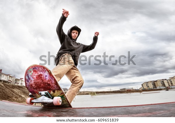 Teen skater in a hoodie\
sweatshirt and jeans slides over a railing on a skateboard in a\
skate park