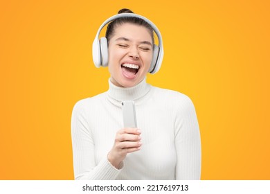 Teen student girl in white turtleneck wearing wireless headphones, singing loud in smartphone as microphone, having fun, isolated on yellow background