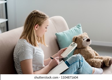 Teen Student Girl Writing In Notebook And Using Calculator While Sitting On Couch