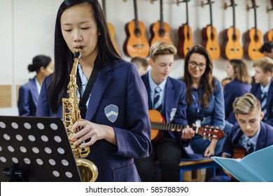 Teen Student Is Playing The Saxophone In Her School Music Lesson. The Rest Of The Class Are In The Background, Out Of Focus.