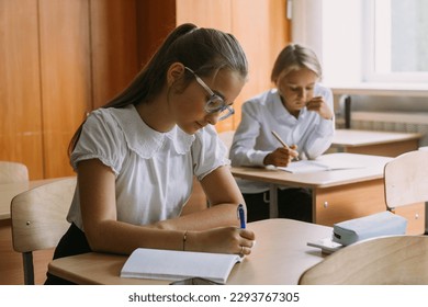 teen students writing in copybook at desk in classroom, looking at camera