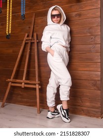  Teen Style. Young Blonde Girl In White Oversize Suit With Hoodie Stands Casual With Hands In Pockets And Smiles Cute In Black Sunglasses Near The Wooden Wall Background. Lifestyle Concept, Free Space