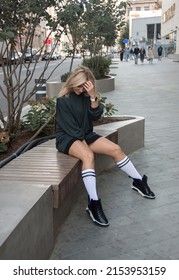 Teen Walk. Glamour Blonde Girl In Black Oversize Dress Is Sitting Casual In White Gaiters On Legs On The Street Bench Background And Smiling Cute With Hand Near Hair . Lifestyle Concept, Free Space