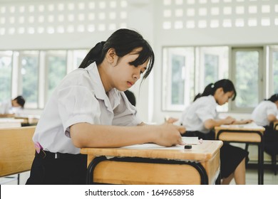 Teen young Asian students group concentrate one's mind writing test in exam on paper answer sheet and sitting on taking final examination ,in rows desk at classroom with Thai student uniform