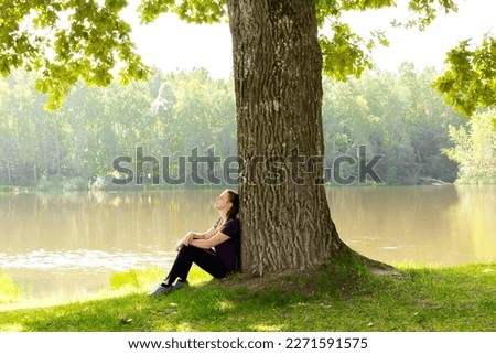 Teen young girl woman sitting on grass under huge high big old oak,trunk tree near lake,river, meditating,relaxing and smiling in National Park.inspiration nature beauty harmony landscape outdoors.