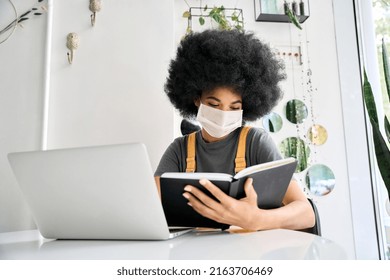 Teenage African American Female College Or University Student With Afro Hair Sitting In Indoor Cafe Learning With Laptop Reading Book Wearing Surgical Face Mask For Covid 19 Prevention.