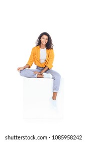 teenage african american student girl sitting on white cube and looking at camera isolated on white