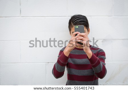Teenage Asian boy standing alone against an urban, white, outdoor background, holding his smartphone up to obscure his face. Teen social concepts: Lost in social media, addicted to social media