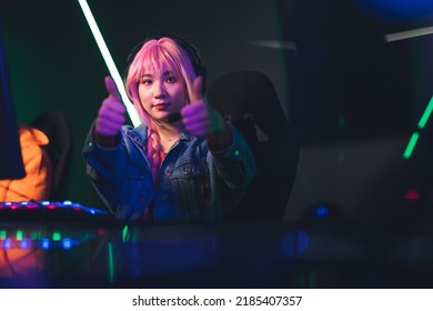 Teenage Asian Pink-haired Girl Sitting In Gaming Chair By A Desk Giving Thumbs-up To The Camera. Gaming Computer Setup. Colored Lights. High Quality Photo