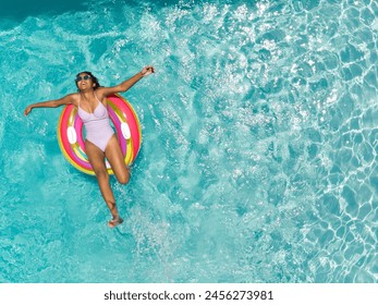 Teenage Biracial girl enjoys a refreshing swim in a pool with copy space on a sunny day. Her relaxed pose on a colorful float suggests a leisurely summer vibe at an outdoor setting. - Powered by Shutterstock