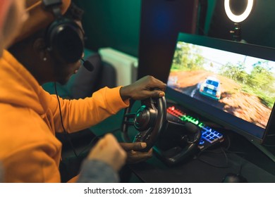 Teenage Bluck Guy Wearing Headset Playing Racing Game With Steearing Wheel In Front Of Big Sceen With Ring Light. Gaming Setup. High Quality Photo