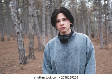 Teenage Boy 16-17 Year Old Wear Sweatshirt And Headphones Posing In Woods Looking At Camera. Sad Serious Guy Outdoors. 