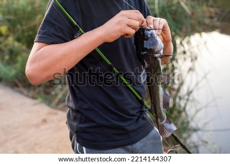 Teenage boy in black t-shirt holding fishing rod and taking bullhead fish off the hook. Blurred lake on the background,