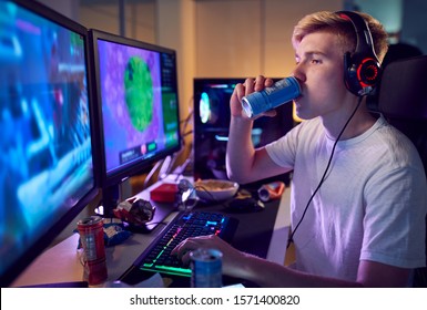 Teenage Boy Drinking Caffeine Energy Drink Gaming At Home Using Dual Computer Screens At Night