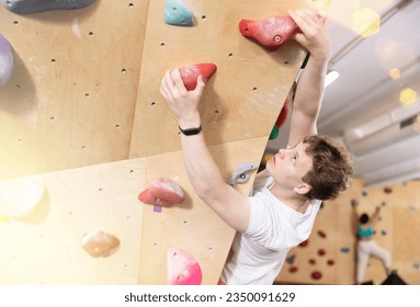 Teenage boy is engaged in fitness clambing in modern equipped gym. Guy visiting sports complex climbs steep artificial wall, top view - Powered by Shutterstock