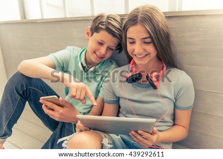 Teenage boy and girl with headphones are using gadgets, talking and smiling while sitting on the floor