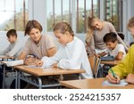 Teenage boy and girl listening to teacher and writing exercises in notebook at lesson in secondary school