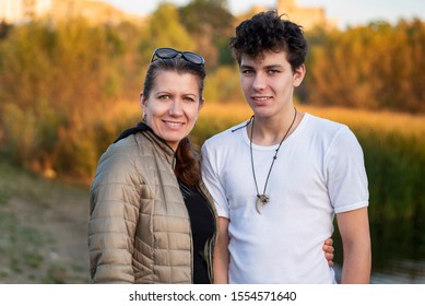 Teenage Boy With His Mom Walking In The Autumn Park And Talking About Life And Having Fun Together. An Authentic Photo Of A Mother And Son. The Woman Gently Hugs Her Almost Adult Son