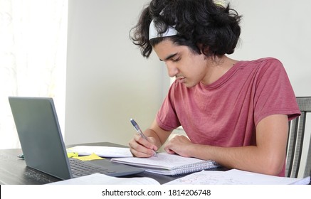 Teenage Boy Of Middle-Eastern Ethnicity Doing Remote High School In Times Of Covid-19, Sitting At Table Taking Notes Off A Laptop.                              