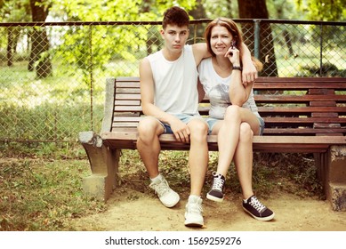 A Teenage Boy With Mother Dressed In T-shirts And Shorts Sitting On A Bench In The Park In Summer And Talking. They Are Trying Together To Overcome The Problems Of Adolescence. Son Hugs Mom