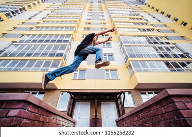 Teenage Boy On A Street In A Big City Next To A High-rise Building Alone. Concept Of A Teenager Parkour Life. Parkour Jump On A High-rise Building Background From The Side Close