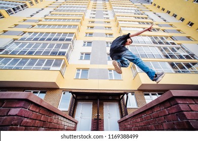 Teenage Boy On A Street In A Big City Next To A High-rise Building Alone. Concept Of A Teenager Parkour Life. Parkour Jump On A High-rise Building Background From The Side Close Up