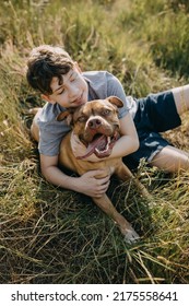 Teenage boy playing with his dog, purebred pit bull, sitting outdoors, in a field. - Shutterstock ID 2175558641