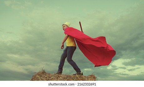 Teenage Boy Plays Knight Against Background Sky. Concept Children Play. Childhood Dream Superhero. Defend Himself With Sword Crown His Head. Superman Winner. Fantasy Childhood Dream. Red Cape Wind