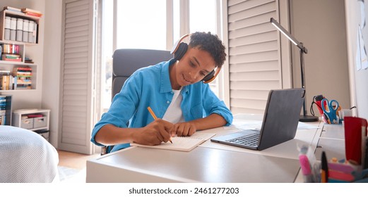 Teenage Boy Sitting At Desk In Bedroom At Home Doing Homework Streaming Music To Headphones - Powered by Shutterstock