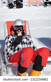 Teenage Boy Smiling, Looking At Camera, Having A Break From Skiing In Mountains And Enjoying A Hot Chocolate Drink In A Recliner Chair On Snow.