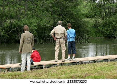 Teenage boy standing on a wooden jetty fishing in a river with a middle aged man standing next to him. Several other people standing nearby, with trees to the rear.