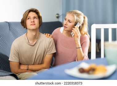 Teenage Boy Suffering From Abdominal Pain Sitting On Sofa At Home, Holding His Stomach And Curving Through Spasms While Concerned Mother Speaking On Phone With Doctor