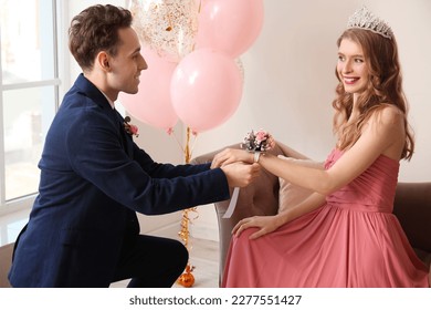 Teenage boy tying corsage around his girlfriend's wrist for prom in room
