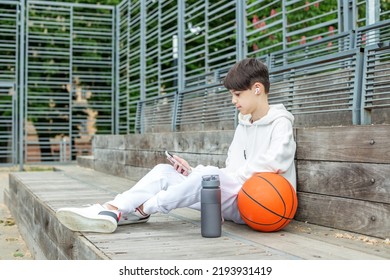 Teenage boy uses smartphone and wireless headphones during break on basketball court. Basketball and reusable water bottle. Concept of sports, school and active lifestyle - Powered by Shutterstock