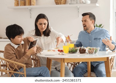 Teenage boy using tablet computer and his angry parents at table during dinner in kitchen. Family problem concept - Powered by Shutterstock