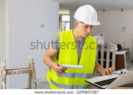 Teenage boy wearing yellow vest and hardhat using laptop and checking documentation in construction site. Young man having architect apprenticeship during part-time work.