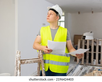 Teenage Boy In Yellow Vest And Hardhat Checking Documents In Construction Site During Workday On Part-time Job.