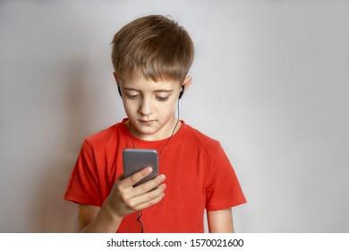 A Teenage Child In A Red T-shirt With Headphones In His Ears Listens To Music From The Phone And Holds It In His Hands. Close-up, Solid Light Background.