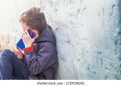 A Teenage European Boy Sits Against The Wall Of The House And Learns About The News On A Blue Smartphone. Difficult Conversation.