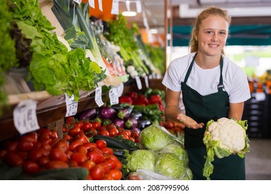 Teenage Female Seller In Uniform Working In Supermarket At Her First Job, Showing Cauliflower