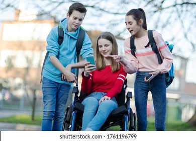Teenage Friends With Girl In Wheelchair Looking At Mobile Phone As They Leave High School