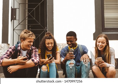 Teenage friends sitting on a pavement holding their mobile phones. Cheerful college boys and girls having fun talking sitting outdoors in a street.