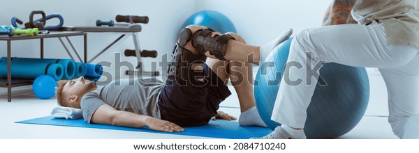 Teenage girl after\
car accident with orthosis on the leg exercises on blue mat in the\
company of\
physiotherapist
