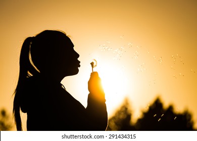 Teenage Girl Backlit By The Sun Blowing A Dandelion Seed Head, Seeds Catching The Sunlight Copy Space To The Right Top Of Image