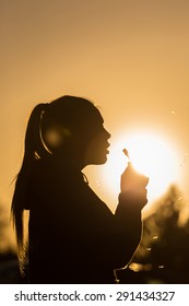 Teenage Girl Backlit By The Sun Blowing A Dandelion Seed Head, Seeds Catching The Sunlight Copy Space To The Right Top Of Image