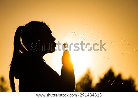 teenage girl backlit by the sun blowing a dandelion seed head, seeds catching the sunlight Copy space to the right top of image