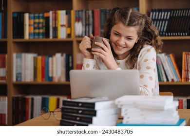 Teenage girl beams with a smile, holding a mug while engaging with her laptop in a library full of books - Powered by Shutterstock