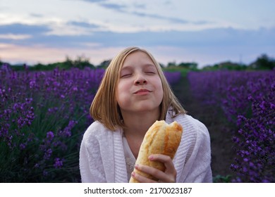 Teenage girl bites fresh French baguette in lavender field. Teenager has a picnic in purple flowers at sunset and enjoys it. Summertime, travel, vacation