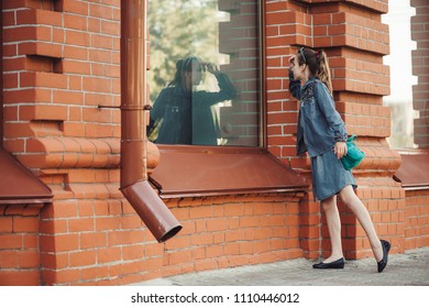 Teenage Girl In Blue Denim Dress Look In Window Of Building From Red (brown) Brick Outdoors. Young Serious Child Piddle Near Shop. Reflex Of Dreamer In Big Window. Girl Wants To Do Shopping.